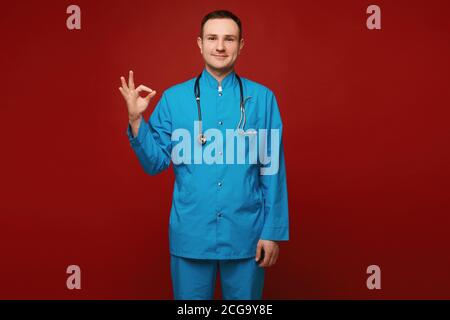 Un giovane in uniforme medica e con uno stetoscopio che mostra un gesto OK e posa su sfondo rosso, isolato. Concetto di assistenza sanitaria e di emergenza Foto Stock