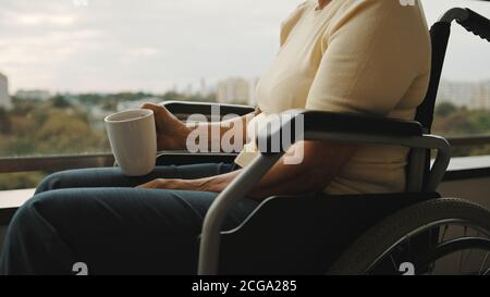 donna anziana in sedia a rotelle sulla terrazza con una tazza di tè o caffè. Nessuna faccia. Foto di alta qualità Foto Stock