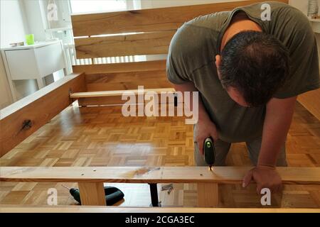 Un uomo che si avvita in travi di legno nel montaggio di un nuovo letto come miglioramento della sua casa. Sta usando un cacciavite a batteria. Foto Stock