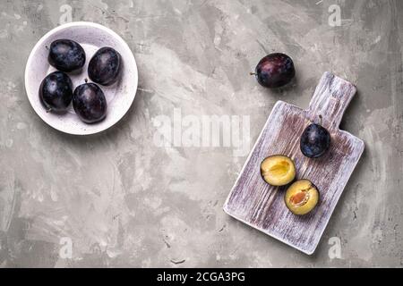 Frutta fresca di prugne matura intera e tagliata in ciotola e su tagliere di legno, fondo di pietra calcestruzzo, spazio di copia vista dall'alto Foto Stock