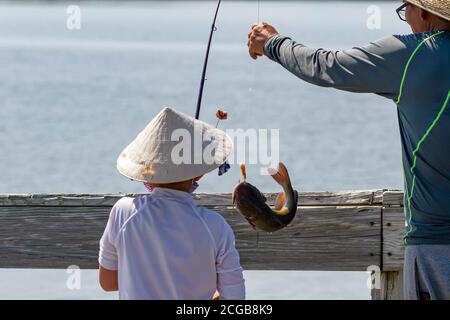 Un ragazzo asiatico che indossa tradizionale bambù cinese cool paglia cappello conico è la pesca con suo padre su un ponte. Il ragazzo ha appena catturato un pesce gatto un Foto Stock