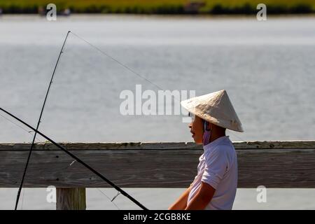 Eastern Neck Island, MD, USA 09/06/2020: Un ragazzo asiatico che indossa un tradizionale cappello di cono di bambù di paglia cinese cool sta tenendo una canna da pesca su un ponte Foto Stock