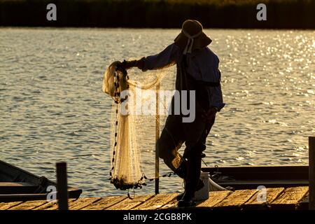 Un pescatore che indossa tute, stivali di plastica e un cappello a secchio sta preparando la sua rete gettata prima di gettarla nella baia di Chesapeake al tramonto. È acceso Foto Stock