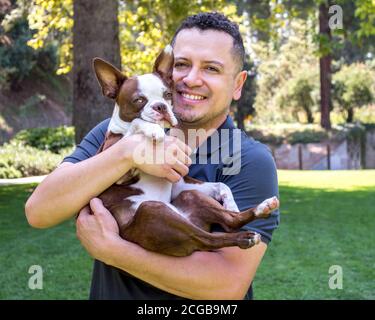 Un giovane ispanico abbracciò un adorabile terrier di Boston cucciolo all'aperto in un parco Foto Stock