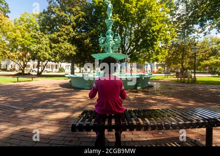 Una bambina che indossa abiti rosa casual è seduta da sola su una panchina di metallo nel Fountain Park di Chestertown, Maryland. Sullo sfondo sfocato Foto Stock