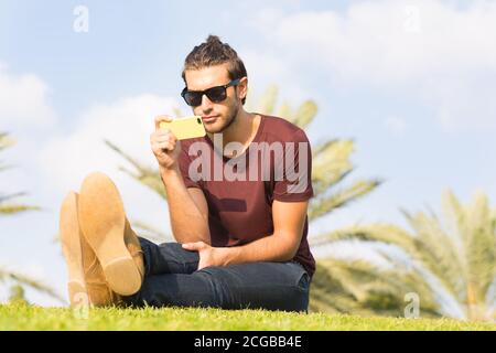 Bell'uomo giovane che usa un telefono cellulare mentre si siede sull'erba nel parco in una soleggiata giornata estiva. Foto Stock