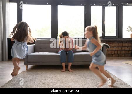 Giovane madre stanca soffre di mal di testa occhi chiusi toccare fronte sedendosi sul divano mentre le sue figlie corrono intorno Foto Stock