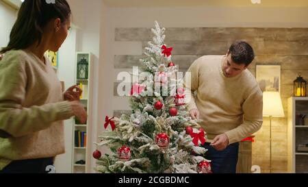 Uomo caucasico e donna felice di decorare il loro albero di natale. Vacanze invernali. Decorazione bello albero di Natale con decorazioni palla di vetro. Moglie e marito in abiti coordinati che aiutano a casa ornata con luci di ghirlanda Foto Stock