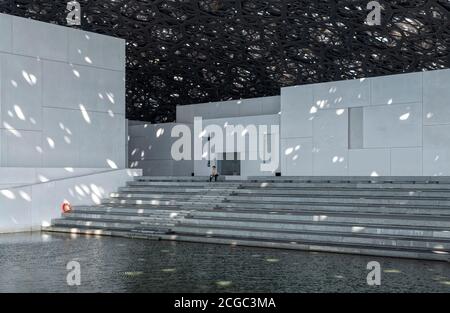 Un'immagine interna del Louvre Abu Dhabi che include un uomo seduto al bordo dell'acqua, la sua luce ad alta tecnologia che filtra dal tetto sopra, e le pareti bianche della facciata circostante di vari spazi espositivi. Aperto a novembre 2017. Foto Stock