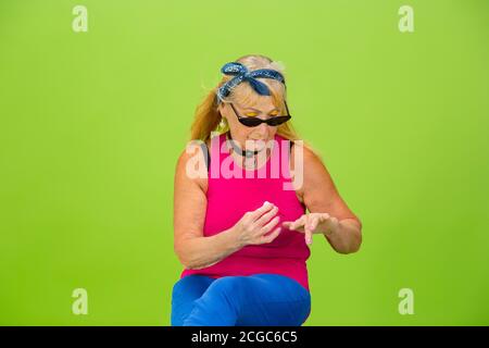 Manicure rossa. Donna anziana in abbigliamento ultra trendy isolato su sfondo verde brillante. Look elegante e alla moda, per sempre giovani. Donna caucasica matura in occhiali da sole, abbigliamento luminoso e sneakers. Foto Stock