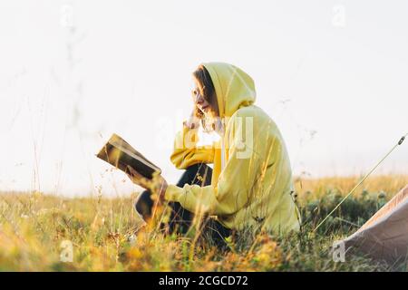 Giovane donna viaggiatore in giallo felpa con cappuccio lettura libro contro tenda. Turismo locale, viaggio nel fine settimana Foto Stock