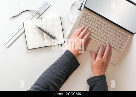 Mani anziane che lavorano su una tastiera per laptop, scrivania bianca da ufficio con notebook e penna, vista dall'alto Foto Stock