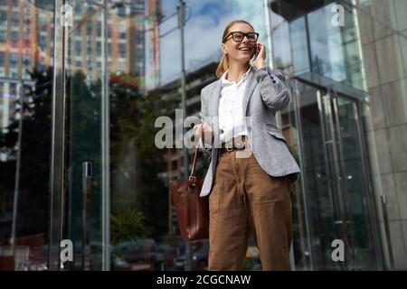 Buona conversazione. Ritratto di felice donna di affari alla moda che indossa occhiali e abbigliamento classico che parla sul telefono cellulare e sorridente mentre si sta in piedi contro Foto Stock