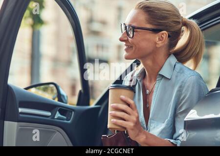 Felice riuscito metà anziana donna di affari che tiene la tazza di carta del caffè mentre esce dalla sua automobile, sta arrivando al lavoro Foto Stock