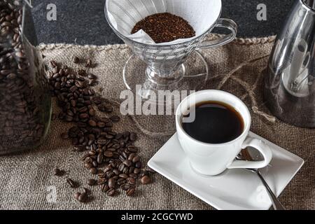Caffè in una tazza bianca su un panno di iuta con caraffa di caffè argento, filtro e chicchi di caffè per la decorazione Foto Stock