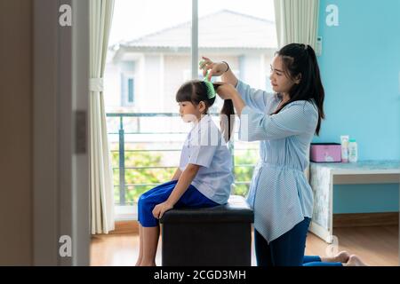 La madre asiatica sta combattendo i capelli della figlia la mattina prima di andare a scuola nel soggiorno a casa. La routine scolastica mattutina per il giorno negli li Foto Stock