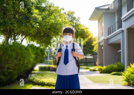 Ritratto di carina ragazza della scuola primaria asiatica in maschera medica di fronte a casa durante la pandemia COVID-19. La routine scolastica mattutina per giorno nella vita g Foto Stock