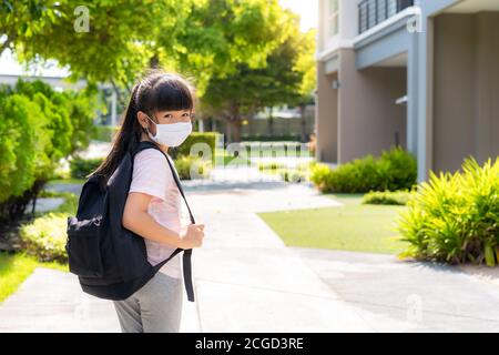 Ritratto di carina ragazza della scuola primaria asiatica in maschera medica di fronte a casa durante la pandemia COVID-19. La routine scolastica mattutina per giorno nella vita g Foto Stock