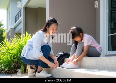 La madre asiatica aiuta la sua figlia studenti primari in uniforme a. indossando la loro scarpa davanti a casa nel la routine scolastica mattutina per il giorno in Foto Stock