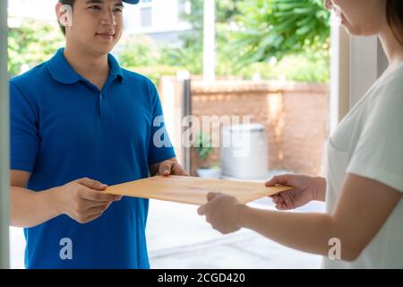 Il giovane uomo di consegna asiatico in sorriso uniforme blu e tenendo la lettera o la busta del documento nella casa anteriore e la donna asiatica che accetta una consegna della busta Foto Stock