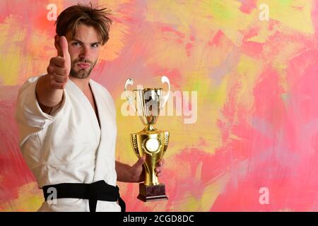 MMA combattente con corpo forte celebra la vittoria. Primo premio e concetto di successo. Uomo con faccia seria su sfondo colorato. Guy posa in bianco kimono mostrando pollici in su tiene premio d'oro Foto Stock