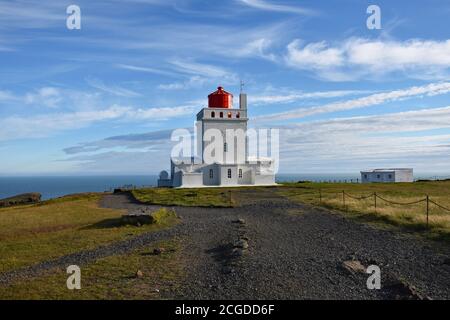 Un sentiero nero conduce verso il faro di Dyrhólaey lungo la costa meridionale dell'Islanda. Un faro bianco con casa lanterna rossa a contrasto. Foto Stock