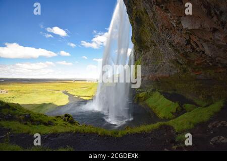 Una vista laterale di Seljalandsfoss in una giornata di sole in Islanda del Sud. La grotta e il sentiero in pietra nera dietro le cascate possono essere visti nella zona ombreggiata. Foto Stock