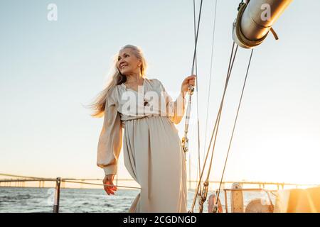 Bella donna senior in abito lungo guardando lontano all'aperto. Donna caucasica matura in piedi su un ponte barca a vela. Foto Stock
