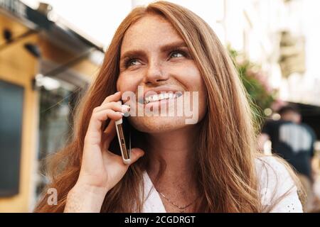 Primo piano di una bella donna sorridente dai capelli rossi seduta al bar all'aperto, che parla sul telefono cellulare Foto Stock