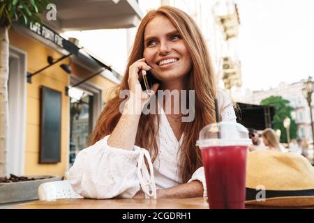 Primo piano di una bella donna sorridente dai capelli rossi seduta al bar all'aperto, che parla sul telefono cellulare Foto Stock