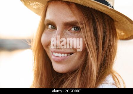 Immagine di gioiosa donna rossa in cappello sorridente e guardando a macchina fotografica mentre camminando sul lungomare Foto Stock