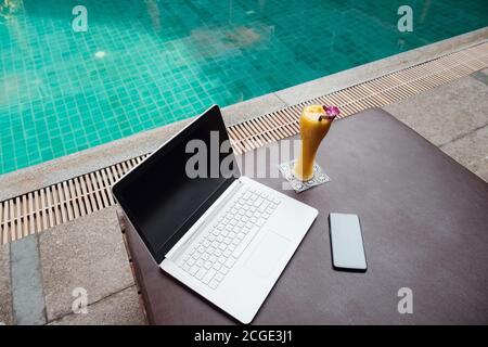 Un portatile bianco, uno smartphone e un frullato di mango su un lettino da sole sullo sfondo della piscina. Un inizio di nuova giornata. Concetto aziendale freelance. Re. Flessibile Foto Stock