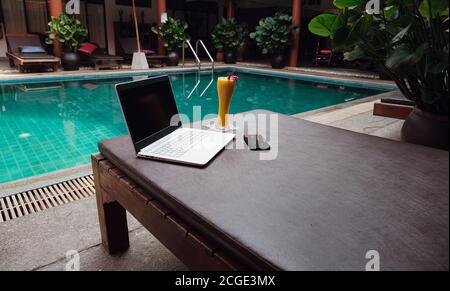 Un portatile bianco, uno smartphone e un frullato di mango su un lettino da sole sullo sfondo della piscina. Un inizio di nuova giornata. Concetto aziendale freelance. Re. Flessibile Foto Stock