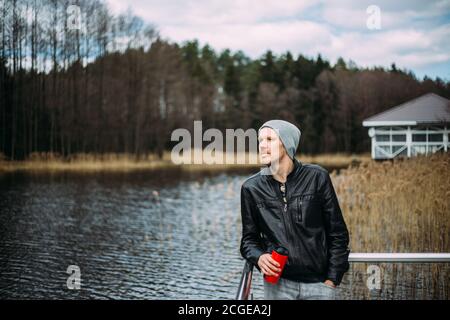 Uomo pusivo in una giacca di pelle e cappello si alza e pensa all'aperto. Buon modello maschile guardando pensivo al lago. Foto Stock