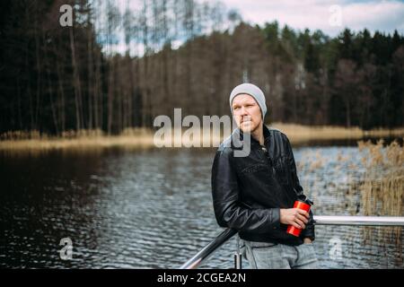 Uomo pusivo in una giacca di pelle e cappello si alza e pensa all'aperto. Buon modello maschile guardando pensivo al lago. Foto Stock