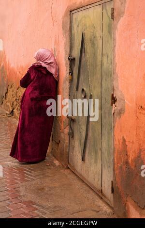 Una donna marocchina in abito tradizionale si appoggia contro un muro intemperie nel souk di Marrakech, Marocco Foto Stock
