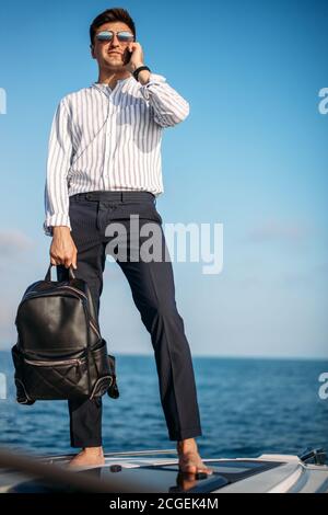 Spazio per il testo. Bel giovanotto con racksack in mano, vestito in abiti da ufficio rilassante e di lavoro su tablet digitale a prua di motoscafo, sul mare Foto Stock