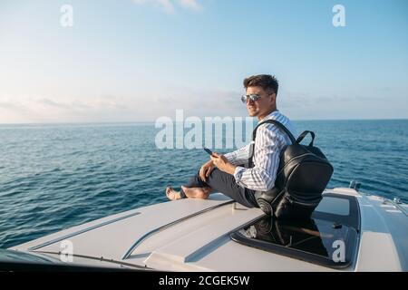 Spazio per il testo. Bel giovanotto con racksack in mano, vestito in abiti da ufficio rilassante e di lavoro su tablet digitale a prua di motoscafo, sul mare Foto Stock