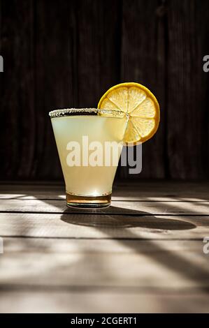 bevanda glassata gialla alcolica estiva in un bicchiere di forma trasparente con zucchero sul bordo e una fetta di limone matura e rotonda su un tavolo di legno, pho Foto Stock