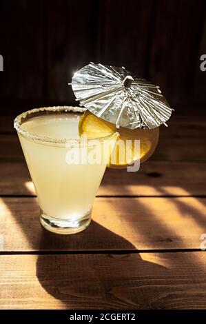 bevanda estiva di colore giallo alcolico in vetro trasparente con zucchero sul bordo, ombrello d'arredo in argento e fetta di lime matura rotonda su tavolo di legno Foto Stock