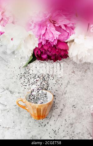 piccola tazza d'oro piena di stelle d'argento e piattino su uno sfondo chiaro con un bouquet di peonie multicolore. Flat Lay, vista dall'alto, Vertica Foto Stock