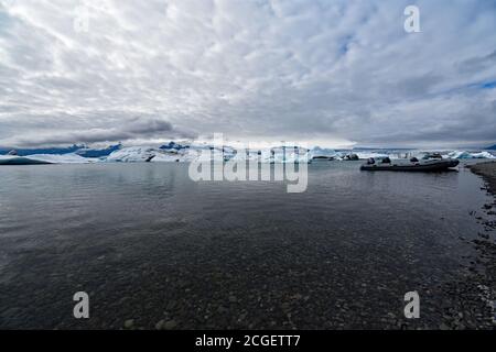 Una barca zodiaca legata sulla riva della laguna glaciale di Jökulsárlón, Islanda del Sud. Gli iceberg galleggiano dietro contro un cielo grigio drammatico in una giornata trascorsa. Foto Stock