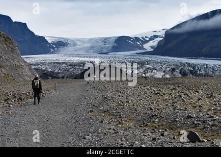 Un escursionista di sesso maschile che indossa un cappello e uno zaino cammina lungo un sentiero di ghiaia verso il ghiacciaio Skaftafellsjokull nel Parco Nazionale di Skaftafell, Islanda. Foto Stock