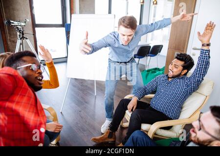 Gruppo di giovani entusiasti che sollevano le mani nello spirito di squadra alto cinque sopra il tavolo, seduti uno di fronte all'altro in uno spazio ben illuminato studio dietro Foto Stock