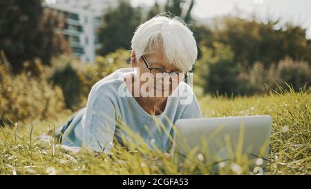 vecchia donna grigia con capelli che usa un computer portatile nella natura mentre si sdraiava sull'erba. Foto di alta qualità Foto Stock