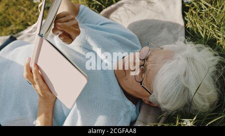 donna grigia anziana che legge un libro mentre riposa sull'erba in natura. Foto di alta qualità Foto Stock