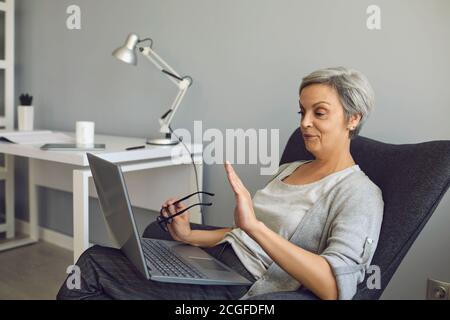 Donna anziana con capelli grigi che ondolano la mano videochiama con il computer portatile a casa. Foto Stock