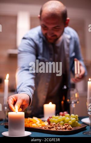 Candela di illuminazione con fiammiferi. Il marito prepara il pasto festivo con cibo sano per la celebrazione dell'anniversario, appuntamento romantico, seduto vicino al tavolo in cucina. Foto Stock