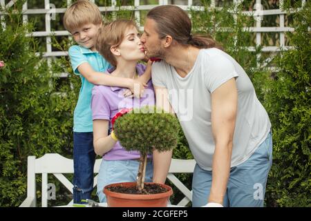 Ritratto di coppia sposata amorevole baciarsi su giardino verde abete-albero sfondo, e felice 6 anni figlio abbracciando Mum e guardando la macchina fotografica. Foto Stock