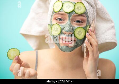 Giovane donna caucasica sana con asciugamano sulla testa, facendo trattamenti e maschera di creta viso con ingredienti organici, chiacchierando su smartphone e mangiando un s. Foto Stock